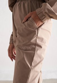 Pantalons de jogging beiges avec taille élastique et poignets élastiques, dotés d'une poche latérale et d'un logo de marque en relief sur la cuisse. Tissu lisse et léger.