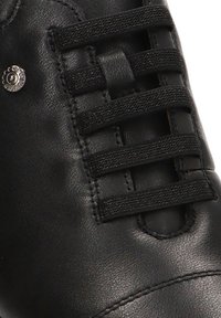Zapato de cuero negro con cordones elásticos, que presenta un acento metálico circular y una textura suave. Enfoque en el sistema de cordones y los detalles de las costuras.