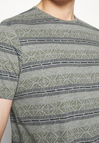 Szara koszulka z krótkim rękawem, zdobiona geometrycznym wzorem zielonych i granatowych pasków, wykonana z miękkiego, teksturowanego materiału.