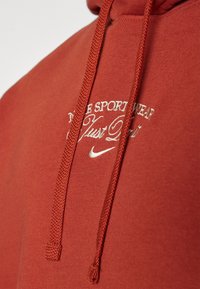 Mörkröd sweatshirt med en mjuk textur, med en dragsko i huva. Broderad vit text som säger "NIKE SPORTSWEAR Just Do It," med en liten logotyp.