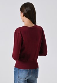 Donna con capelli scuri che indossa orecchini dorati, un maglione bordeaux aderente e jeans blu, voltata di spalle su uno sfondo semplice.