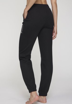 Woman barefoot wearing black jogger pants with elastic cuffs and side pockets, standing against plain background, back view.