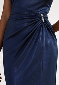 Σκούρο μπλε φόρεμα με σχέδιο ρυτιδώσεων και μεταλλική χρυσή λεπτομέρεια στη μέση, κατασκευασμένο από λαμπερό, υφασμάτινο ύφασμα με υφή.