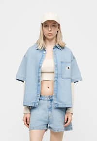 Άτομο που φοράει ανοιχτό μπλε τζιν πουκάμισο με κοντά μανίκια και σορτς πάνω από μπεζ κοντό τοπ και μακριά μανίκια, με λευκό καπέλο.