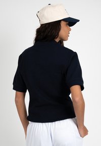 Femme de dos portant un t-shirt noir à manches courtes, un short en mesh blanc et une casquette blanche et marine.