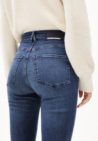 Ylhäältä korkeavyötäröiset siniset farkut, joissa on kaksi takataskua, musta logopläkki vyötäröllä ja tiukka istuvuus lahkeissa.