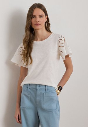 Femme aux longs cheveux bruns portant un chemisier blanc à manches volantées en dentelle et un pantalon taille haute bleu clair, debout contre un mur uni.