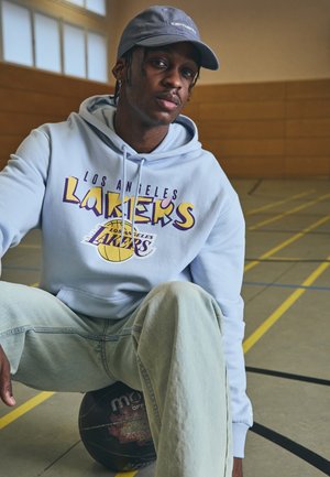 Light blue hoodie with "Los Angeles Lakers" graphic, paired with light denim jeans. A basketball sits beneath him on a gym floor.