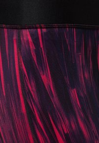 Legginsy mają czarny pas z teksturowanym, abstrakcyjnym wzorem w kolorze czerwonym i fioletowym. Gładka tkanina z kontrastującymi detalami szwów.