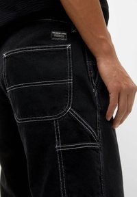 Primer plano de un pantalón vaquero negro con costuras blancas y múltiples bolsillos de utilidad, mostrando la mano derecha de una persona descansando junto al muslo.