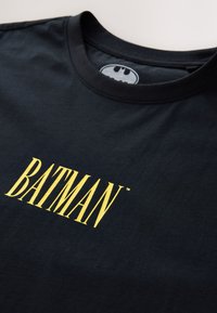 Camiseta negra de algodón que presenta un gran gráfico de texto amarillo "BATMAN" en el frente. Diseño de cuello redondo con una textura suave.