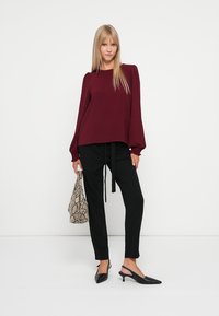 Blusa de manga longa cor borgonha, de corte solto, combinada com calças pretas de corte tapered e sapatos de salto pretos tipo slingback. Segurando uma mala texturizada com padrão de cobra.