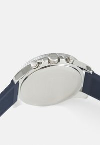 Reloj de acero inoxidable plateado con una caja redonda lisa, correa de goma azul marina y corona texturizada. Las características incluyen dos botones de presión y una parte trasera sin marcar.