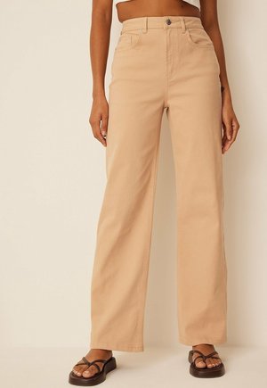 Beige bukser med høj talje og brede ben, kombineret med mørke plateau-sandaler, vist fra midten af torsoen til fødderne mod en ensfarvet baggrund.