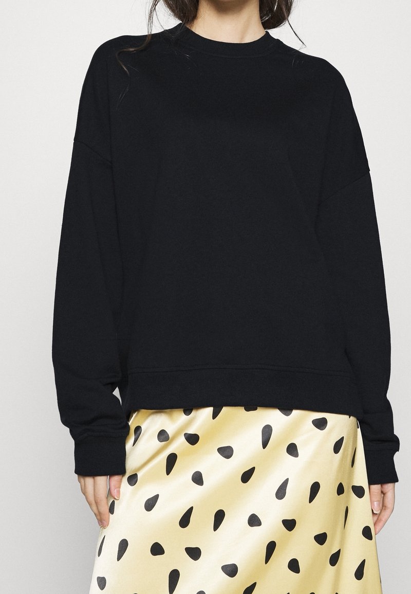 Femme portant un pull noir à manches longues et une jupe jaune brillante avec des taches noires irrégulières.
