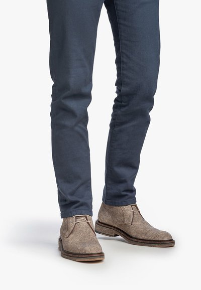 Man wearing slim dark blue pants and light brown suede lace-up ankle boots standing on white background.