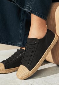 Mustad mustad kingad naturaalsest jute materjalist toe cap'i ja äärisega, millel on nööbid ja tekstureeritud tald, esitatud heleda taustaga.