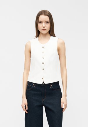 Jeune femme debout face à l'avant, portant un haut blanc sans manches à boutons et un jean bleu foncé taille haute, sur un fond blanc uni.