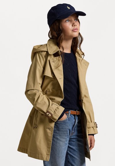 Beige trenchcoat med dobbeltknappet design, knapper og belte i midjen, kombinert med en marineblå caps og en marineblå genser.