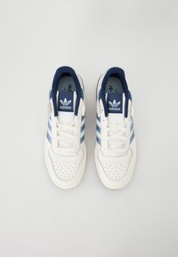 Biele tenisky s námorníckymi a svetlomodrými pásmi, perforovanou špičkou a šnúrkami. Obsahujú modrý polstrovaný golier a logo Adidas na jazyku.