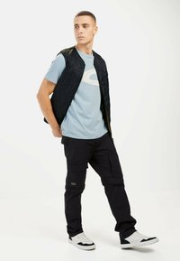 Sort quiltet vest over en lyseblå t-shirt med et logo, parret med mørke cargo-bukser og hvide sneakers. Enkel, funktionel design.