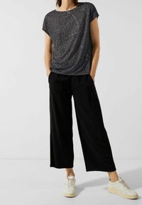 Černé volné široké kalhoty kombinované s krátkým topem s tmavým vzorem a jemnými ozdobami. Celkový vzhled doplňují bílé tenisky.