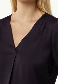 Blouse violet profond avec un décolleté en V, des plis à l'avant, fabriquée en tissu lisse. Le modèle présente des manches courtes.