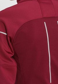 Bordowa kurtka sportowa z gładkiego materiału, z wysokim kołnierzem i odblaskowymi białymi paskami wzdłuż pleców i ramion dla lepszej widoczności.