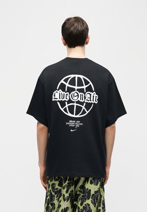 Man draagt een zwart T-shirt met een witte wereldbol en de tekst "Live On Air" en camo-gepatroonde groene en zwarte shorts, met zijn rug naar de kijker gekeerd.