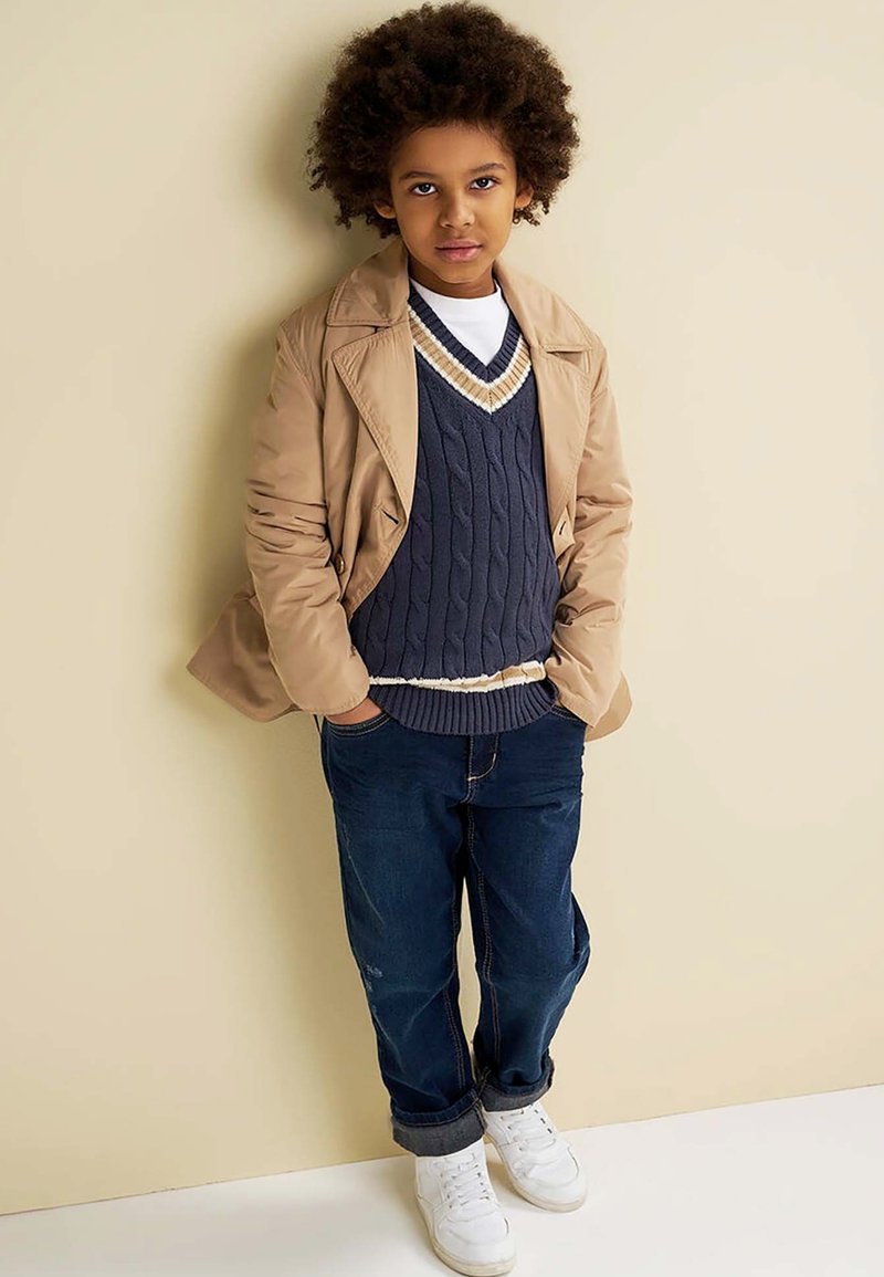Beige trenchcoat, marineblauwe kabeltrui met crèmekleurige accenten, blauwe jeans en witte sneakers. Kind met krullend haar staat tegen een beige achtergrond.