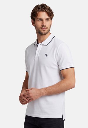 Mann mit kurzen lockigen Haaren, der ein weißes Polohemd mit schwarzem Besatz und einem kleinen gestickten Logo auf der Brust trägt, steht vor einfachem Hintergrund.