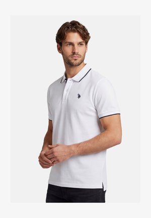 Mann mit kurzen lockigen Haaren, der ein weißes Polohemd mit schwarzem Besatz und einem kleinen gestickten Logo auf der Brust trägt, steht vor einfachem Hintergrund.