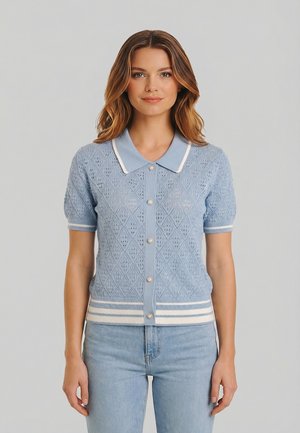 Giovane donna che indossa un cardigan a maniche corte di maglia azzurra con motivo a rombi, bottoni a perla e colletto e orlo a righe, abbinato a jeans blu.