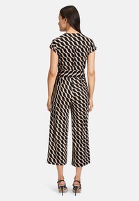 Jumpsuit a motivo ondulato nero e crema con corpino aderente, maniche a cappuccio, pantaloni a gamba larga e dettaglio della vita annodata. Vista del modello in piedi da dietro.