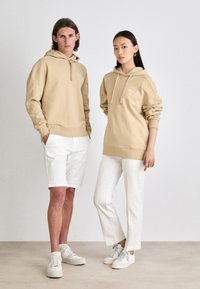 Beiga huvtröjor med snörning, bärs av två modeller. En modellpar har vita shorts och sneakers; det andra par har vita raka byxor och sneakers.