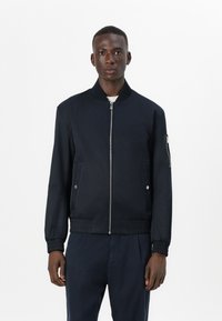 Veste bomber en tissu bleu marine avec fermeture éclair à l'avant, poches latérales et poignets élastiques. Design simple avec une texture lisse et peu de détails.