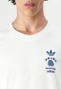 White cotton t-shirt with crew neck, featuring blue graphic print of an Adidas logo and floral design on the left chest area.