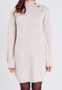 Abito maglione a costine di un beige chiaro con collo alto e maniche lunghe. Presenta bottoni decorativi su una spalla e una consistenza morbida.