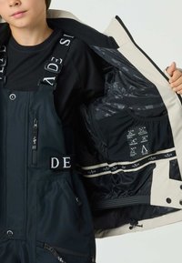 Black and navy snow bib with adjustable straps, zippers, and pockets. Interior features a smooth, black lining with care instructions.