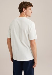 Camiseta blanca de manga corta con patrón texturizado, escote redondeado y un ajuste relajado, vista desde la espalda sobre un fondo neutro.