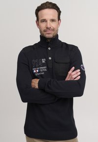 Mornarski pleten pulover s visokim ovratnikom, zapenjanjem spredaj, žepom na prsih in vezenimi detajli. Ima rebraste manšete in rob.