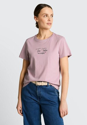 Femme portant un t-shirt Tom Tailor violet clair rentré dans un jean bleu avec une ceinture beige, regardant vers sa droite sur un fond uni.
