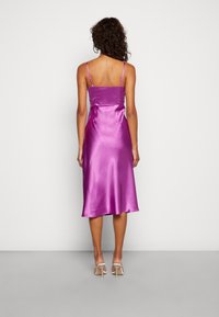 Femme aux cheveux bouclés portant une robe midi en satin violet brillant à fines bretelles, debout face à un mur gris clair uni, chaussée de talons hauts transparents.