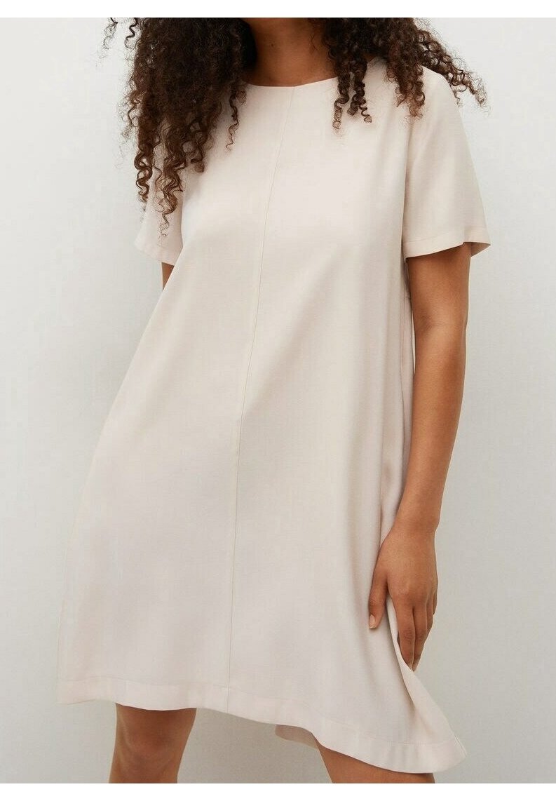 Femme portant une robe beige à manches courtes et coupe ample, debout devant un fond clair uni.
