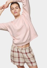 Lichtroze sweatshirt met lange mouwen en een cropped ontwerp; gecombineerd met beige geruite shorts met rode en bruine lijnen, elastische tailleband.