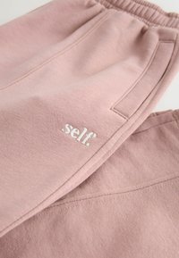 Lyserøde sweatpants lavet af blødt materiale med elastisk linning, med et hvidt broderet logo der står "self." og sidelommer.