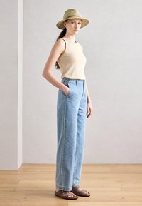 Beige ärmlös topp, ljusblå högmidjade jeans med sidofickor och bruna sandaler med remmar; modell som bär en halmhatt.