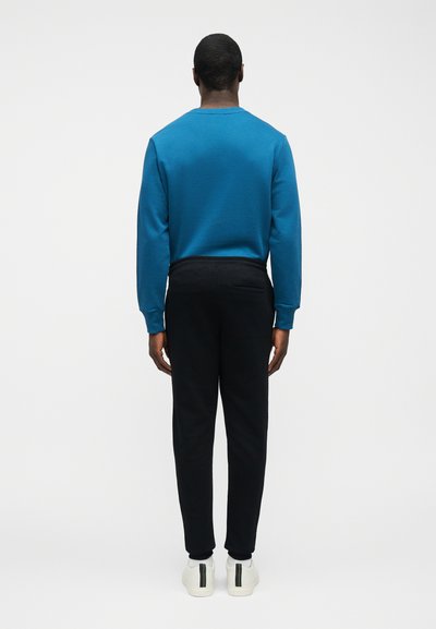 Άνδρας με γυρισμένη πλάτη φοράει μπλε πουκάμισο μακρυμάνικο, μαύρο παντελόνι με τσέπη στο πίσω μέρος και λευκά αθλητικά παπούτσια με μαύρες και πράσινες ρίγες.