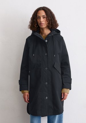 Zwarte waterdichte jas met een capuchon, voorzien van een knoopsluiting, twee voorkruiszakken en een relaxte pasvorm. Gekozen over een mosterdkleurige trui.