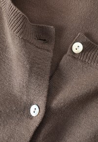 Bruine gebreide cardigan met een geribbelde kraag en twee witte knopen. Soepele textuur met een subtiele glans, met een knoopsgat bij de kraag.